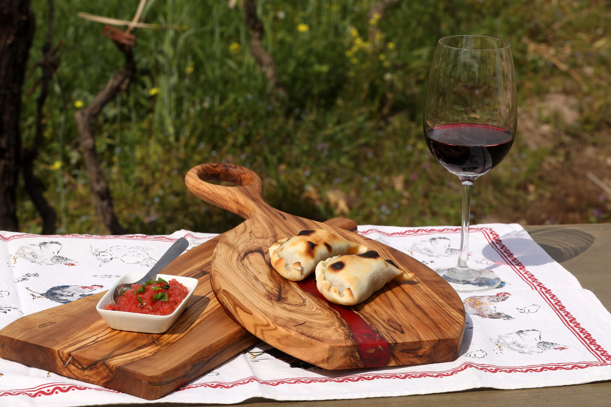 Empanadas caseras servidas sobre una tabla de madera, acompañadas por una salsa y una copa de vino, en una mesa al aire libre.