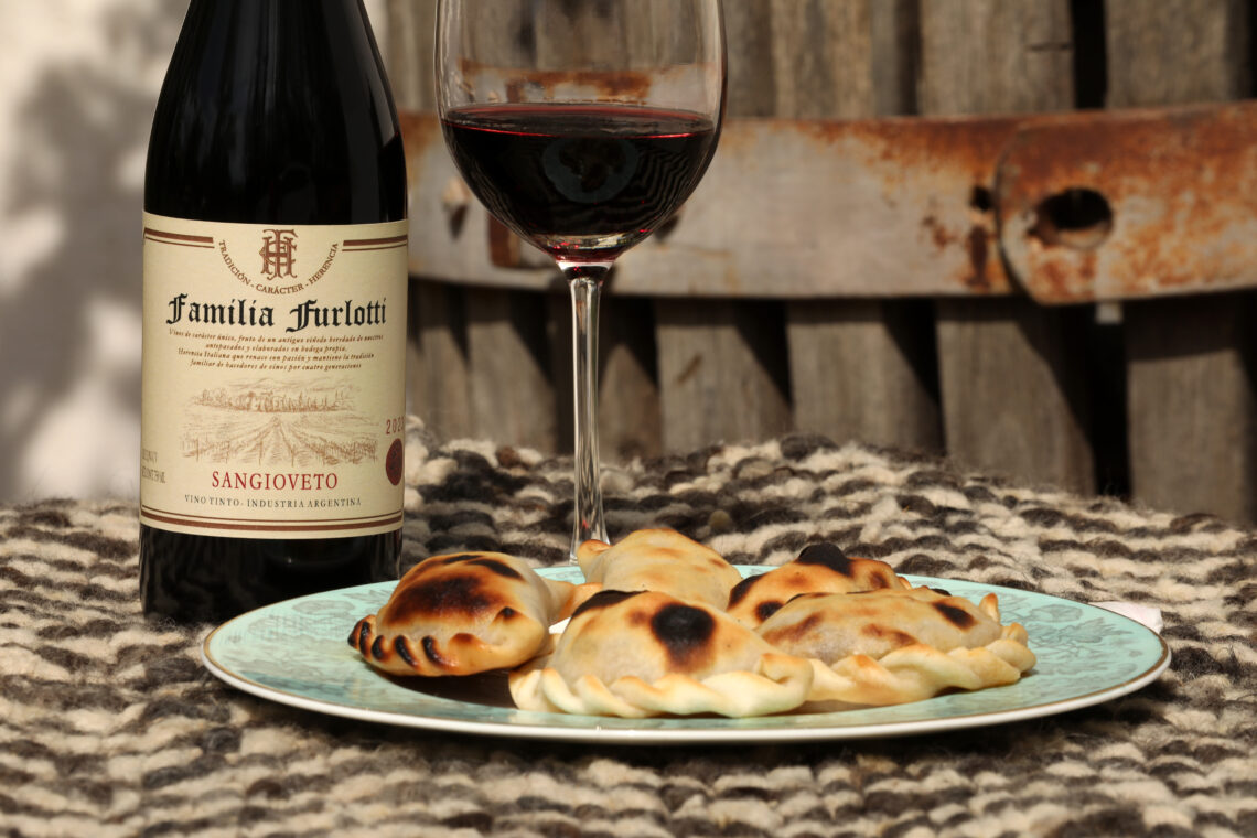 Empanadas caseras servidas en un plato junto a una botella y una copa de vino, sobre una mesa al aire libre, evocando rituales heredados, encuentro y continuidad entre generaciones.
