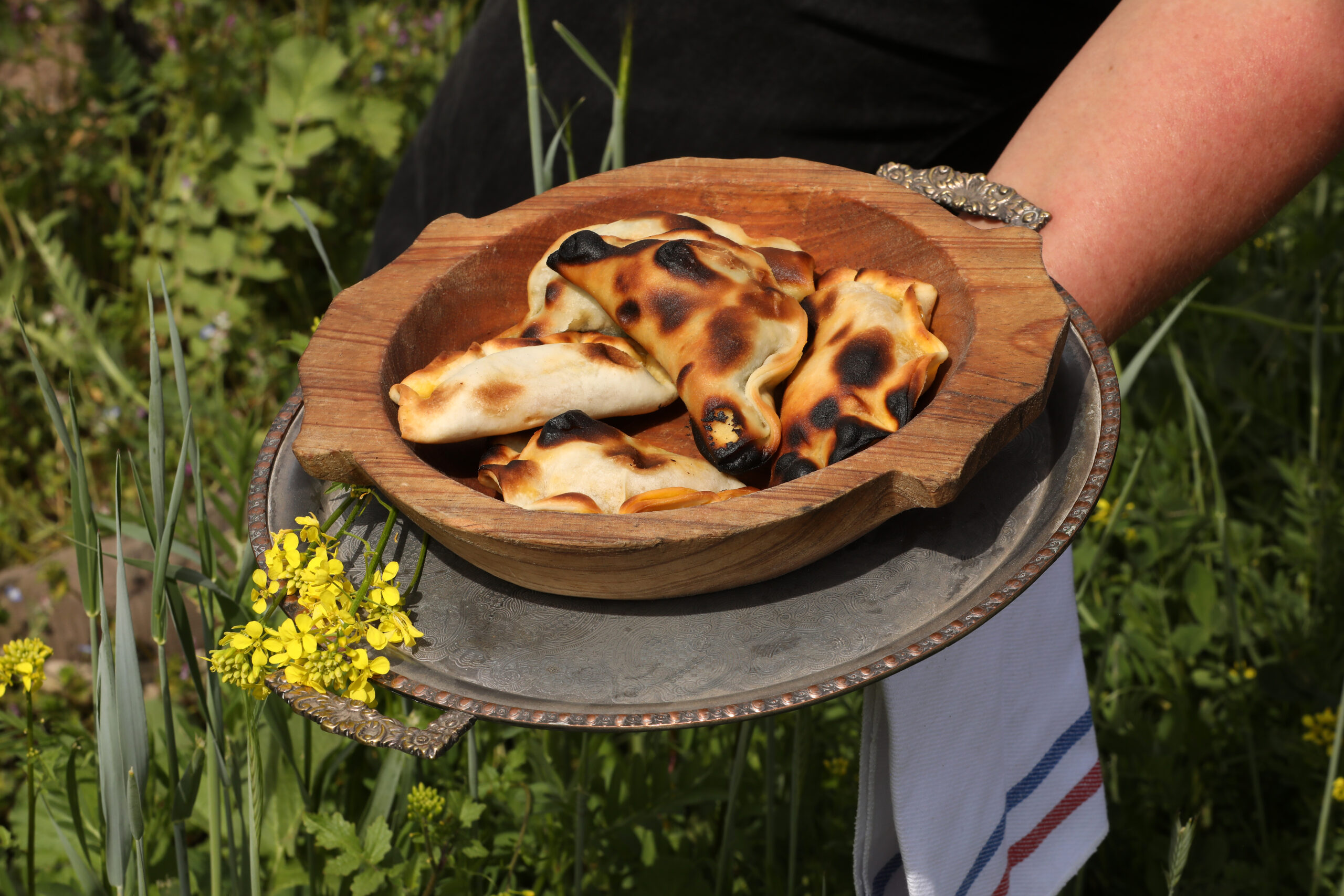 Empanadas caseras doradas servidas en una fuente de madera, presentadas al aire libre entre vegetación, como gesto de cocina tradicional y mesa compartida.