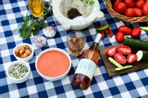 A bottle of Familia Furlotti rosé alongside a bowl of gazpacho on a blue-and-white checkered tablecloth. Surrounding it are fresh ingredients—tomatoes, cucumber, garlic, herbs, olive oil, and chili peppers—along with small bowls of croutons and finely chopped vegetables, creating a fresh, summery scene.