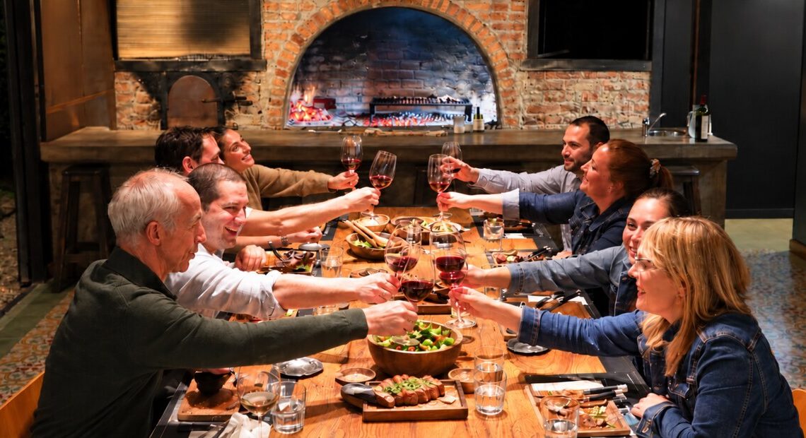 Brindis con vino alrededor de la mesa larga, con el asador cocinando al fondo, en Finca Adalgisa