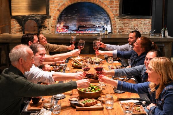 Brindis con vino alrededor de la mesa larga, con el asador cocinando al fondo, en Finca Adalgisa